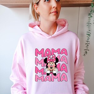 K&ouml;nnte beinhalten: Hellrosa Kapuzenpullover mit dem Wort "MAMA" in rosa Blockbuchstaben, viermal wiederholt. Eine Cartoon-Grafik von Minnie Mouse befindet sich unter dem Text. Die Figur tr&auml;gt eine Sonnenbrille und eine rosa Schleife.