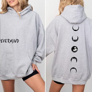May include: A light gray hoodie with the word "NEVERMIND" printed on the front. The back of the hoodie features a black graphic of the phases of the moon. The hoodie has a hood and a front pocket.