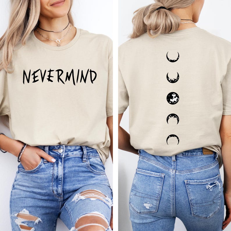 May include: A beige t-shirt with the word "NEVERMIND" in black lettering on the front. The back of the shirt features a black crescent moon design showing the phases of the moon. The shirt is paired with blue jeans.