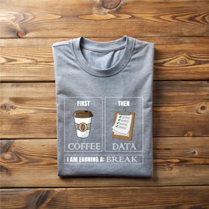 May include: A gray t-shirt with a graphic that reads "First Coffee, Then Data, I Am Earning A: Break." The graphic features a coffee cup and a clipboard. The shirt is folded on a wooden surface.