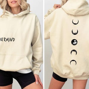 May include: Cream-colored hoodie with the word "NEVERMIND" printed in black on the front. The back features a black graphic of moon phases. The hoodie has a hood and a front pocket.