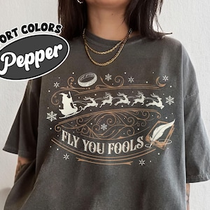 May include: Dark grey Comfort Colors t-shirt with a graphic featuring a sleigh pulled by reindeer, snowflakes, and the text "FLY YOU FOOLS". The shirt has a relaxed fit and a vintage look.