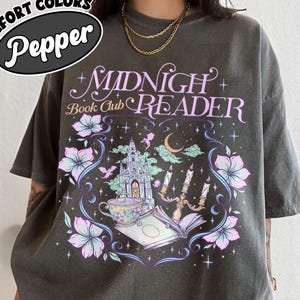 May include: Dark grey t-shirt with the text "Midnight Book Club Reader" in a whimsical design. The graphic includes a castle, flowers, a teacup, and candles. The shirt also has the text "Comfort Colors Pepper" in a circular design.