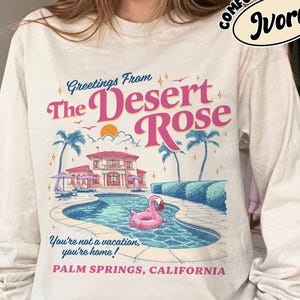 May include: Ivory long-sleeve shirt with a graphic featuring a pool scene with a pink flamingo float, palm trees, and a house. The text reads "Greetings From The Desert Rose" and "Palm Springs, California."