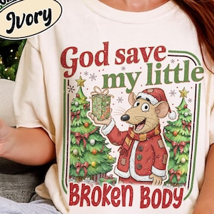 May include: Ivory-colored t-shirt with a Christmas-themed graphic. The graphic features a cartoon rat in a Santa hat and coat, holding a gift box. Text on the shirt reads "God save my little BROKEN BODY". The shirt also has the words "COMFORT COLORS" and "Ivory" on the top left.