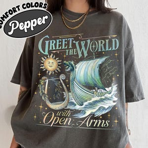 May include: Dark grey t-shirt with a graphic design featuring a Viking ship sailing on waves, a sun, and the text "Greet the World with Open Arms." The shirt has the Comfort Colors Pepper logo.