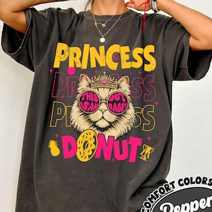 May include: Dark gray t-shirt with a graphic design featuring a cat wearing sunglasses and a crown, with the words "PRINCESS" and "DONUT" in yellow and pink. The shirt also has the text "THIS OUT RAGE".