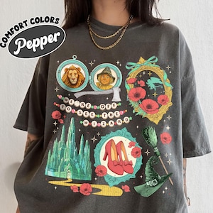 May include: Dark gray t-shirt featuring illustrations from The Wizard of Oz. The design includes portraits of the Lion and Scarecrow, the Emerald City, ruby slippers, and the words "We're off to see the Wizard."