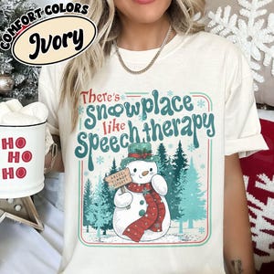 May include: Ivory-colored t-shirt with a snowman graphic and the text "There's snowplace like speech therapy." The snowman wears a green hat, red scarf, and holds a sign. The shirt is part of the Comfort Colors collection.