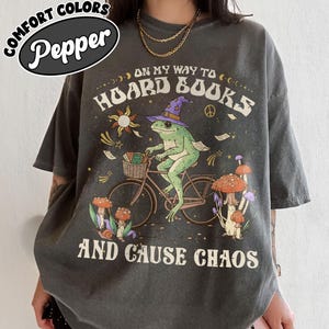 May include: Dark grey t-shirt with a whimsical graphic of a frog riding a bicycle, wearing a wizard hat, and carrying books. Text reads "ON MY WAY TO HOARD BOOKS AND CAUSE CHAOS." The shirt has a Comfort Colors label.