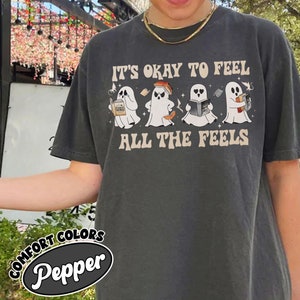 May include: A dark gray t-shirt with the text "It's okay to feel all the feels" and a line of four ghosts with different expressions. Each ghost is holding a book or a cup of coffee.