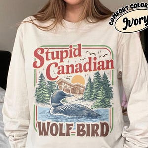 May include: Ivory long-sleeve shirt with a graphic that reads "Stupid Canadian" in red and green. The graphic features a loon on water, trees, and a building. The words "WOLF BIRD" are below the graphic.