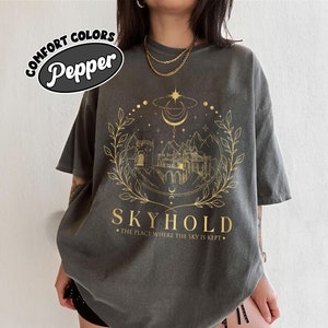 May include: A dark gray t-shirt with a gold graphic of a castle, moon, and stars. The text "Skyhold" and "The place where the sky is kept" is printed below the graphic.