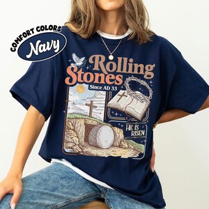 May include: Navy blue t-shirt with a graphic featuring the text "Rolling Stones Since AD 33" and an illustration of a religious scene with a tomb, cross, and open book. The shirt has a white collar.