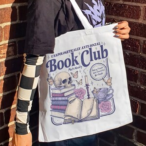 May include: White tote bag with a graphic design featuring a skull, books, butterflies, and a teacup. The text reads "UNAPOLOGETICALLY ANTI-SOCIAL Book Club" and "Making Friends With Characters, Not People."