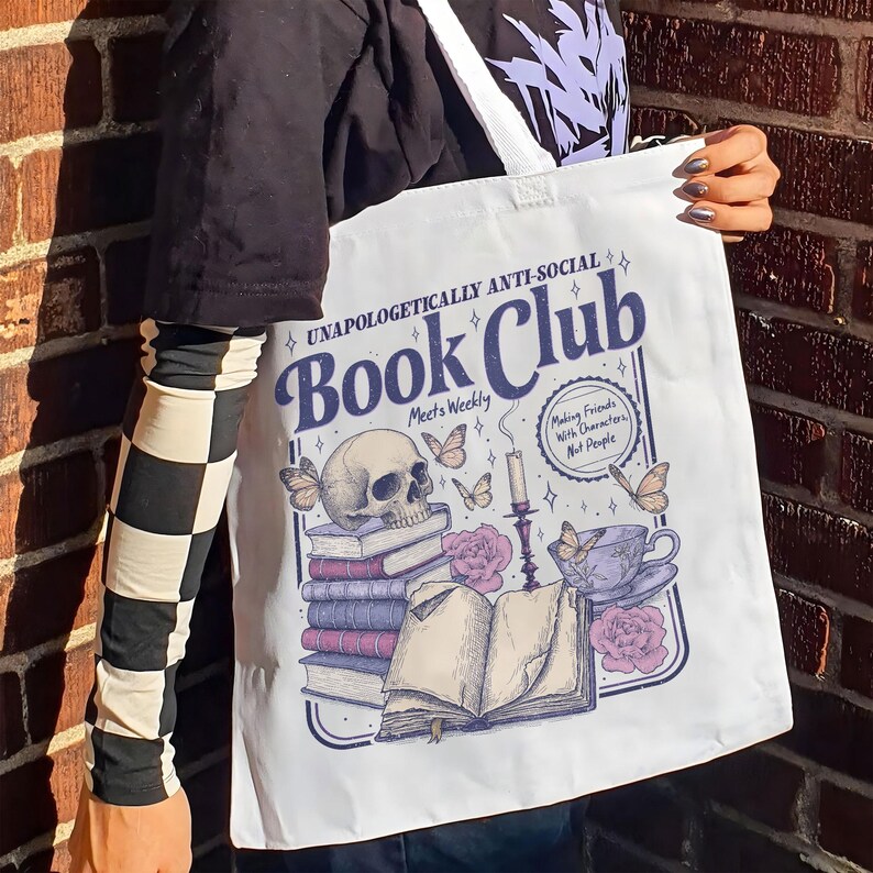 May include: White tote bag with a graphic design featuring a skull, books, butterflies, and a teacup. The text reads "UNAPOLOGETICALLY ANTI-SOCIAL Book Club" and "Making Friends With Characters, Not People."