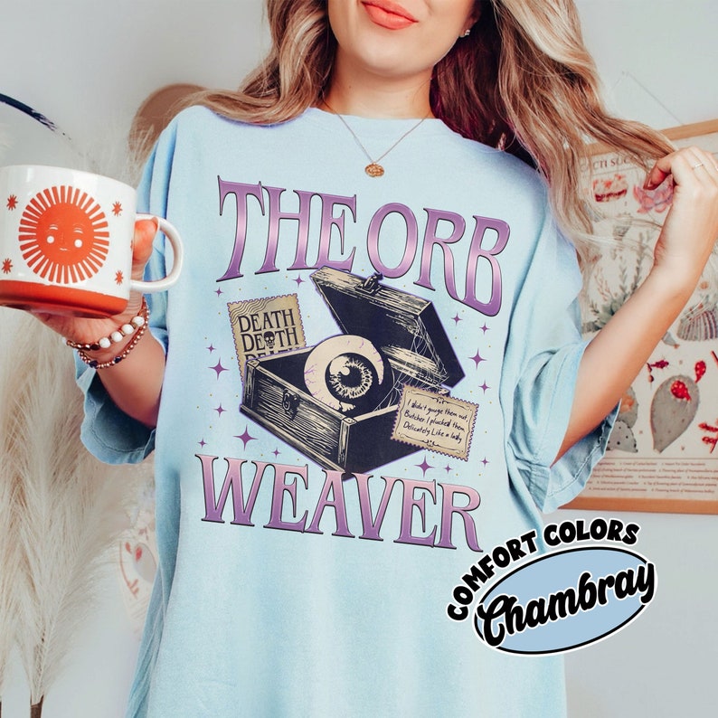 May include: A light blue t-shirt with a graphic of a vintage-style wooden box with an eye inside. The box has a label that says "Death Depth" and the text "The Orb Weaver" is printed in purple below the box. The shirt also has a label that says "Comfort Colors Chambray".