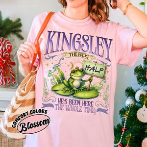 Puede incluir: Camiseta rosa claro con la inscripci&oacute;n "KINGSLEY THE FROG" en morado. La camiseta presenta una rana de dibujos animados con una corona y un cartel que dice "HALP". La camiseta tambi&eacute;n tiene el texto "HE'S BEEN HERE THE WHOLE TIME".
