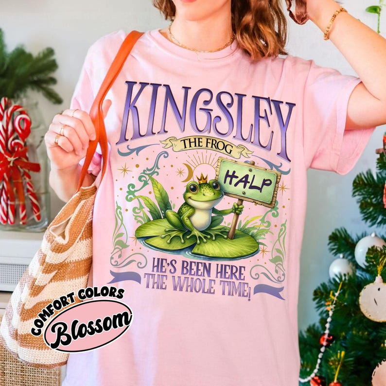 Puede incluir: Camiseta rosa claro con la inscripci&oacute;n "KINGSLEY THE FROG" en morado. La camiseta presenta una rana de dibujos animados con una corona y un cartel que dice "HALP". La camiseta tambi&eacute;n tiene el texto "HE'S BEEN HERE THE WHOLE TIME".