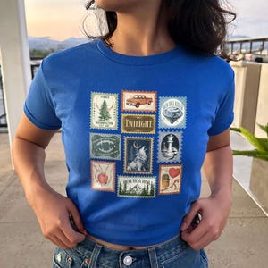 May include: Royal blue t-shirt featuring a collection of postage stamp designs. The stamps depict various images, including a truck, a wolf, a lighthouse, and the word "Twilight". The shirt is a crew neck style.
