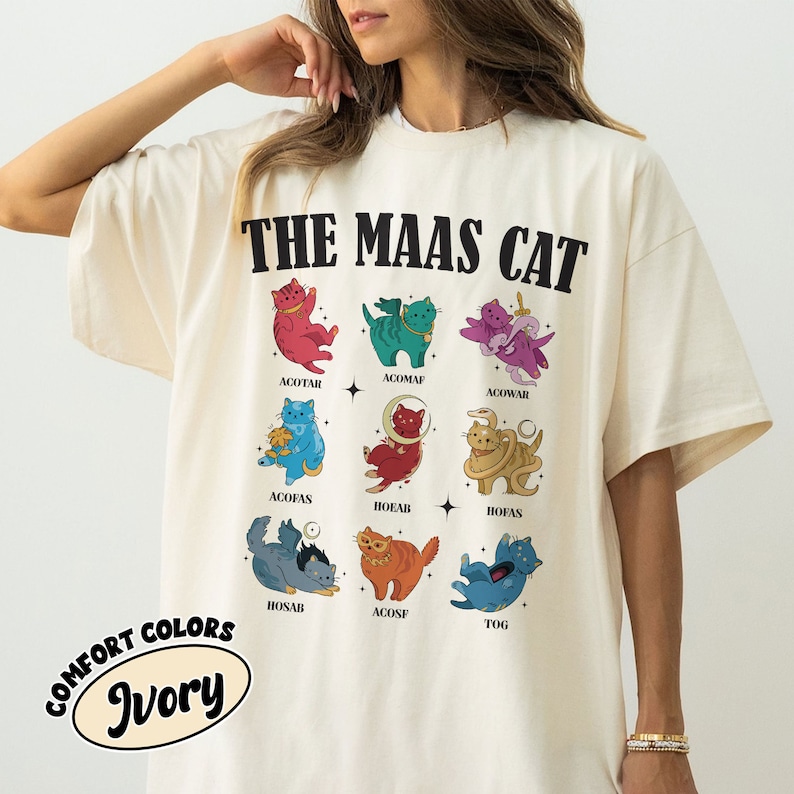 Puede incluir: Una camiseta blanca con el texto "THE MAAS CAT" impreso en negro. La camiseta presenta una cuadr&iacute;cula de nueve ilustraciones de gatos coloridos, cada uno con un dise&ntilde;o &uacute;nico y un nombre escrito debajo.