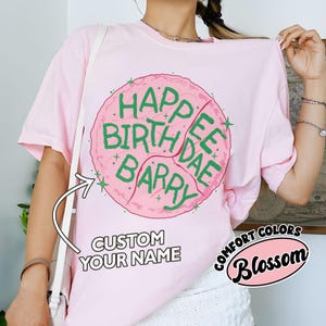 May include: A light pink t-shirt with a large pink graphic that says "Happee Birthdae Barry" in green lettering. The shirt also has the text "Custom Your Name" and "Comfort Colors Blossom".