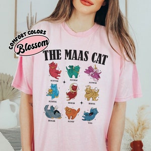 Puede incluir: Camiseta rosa con un gr&aacute;fico de nueve gatos en diferentes colores y poses. El texto "THE MAAS CAT" est&aacute; impreso en negro sobre los gatos. El texto "COMFORT COLORS Blossom" est&aacute; impreso en un c&iacute;rculo en la esquina superior izquierda.