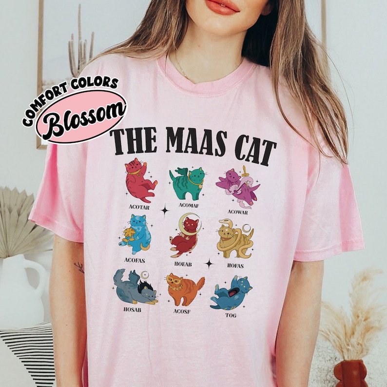 Puede incluir: Camiseta rosa con un gr&aacute;fico de nueve gatos en diferentes colores y poses. El texto "THE MAAS CAT" est&aacute; impreso en negro sobre los gatos. El texto "COMFORT COLORS Blossom" est&aacute; impreso en un c&iacute;rculo en la esquina superior izquierda.