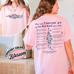 May include: A light pink t-shirt with a floral and sword design on the front and back. The back of the shirt features text that reads "They said 'I love you' but Kai Azer once said..." and a list of quotes. The shirt also has the text "The Powerless Trilogy by Lauren Roberts."