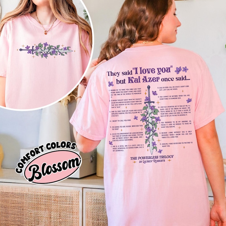 May include: A light pink t-shirt with a floral and sword design on the front and back. The back of the shirt features text that reads "They said 'I love you' but Kai Azer once said..." and a list of quotes. The shirt also has the text "The Powerless Trilogy by Lauren Roberts."