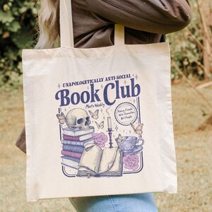 May include: Cream-colored tote bag with a graphic design featuring a skull, books, butterflies, and a teacup. The text reads "UNAPOLOGETICALLY ANTI-SOCIAL Book Club Meets Weekly" and "Making Friends With Characters, Not People."