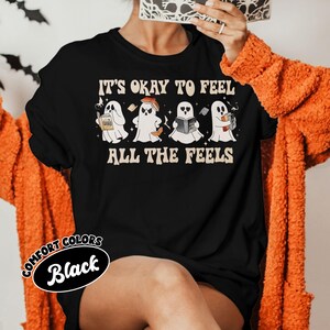 May include: Black t-shirt with a graphic of four ghosts reading books. The text "It's okay to feel all the feels" is printed on the shirt.
