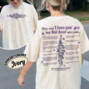 May include: Ivory-colored Comfort Colors t-shirt featuring a floral and sword design with text. The back of the shirt has text from "The Powerless Trilogy" by Lauren Roberts. The shirt is a relaxed fit.