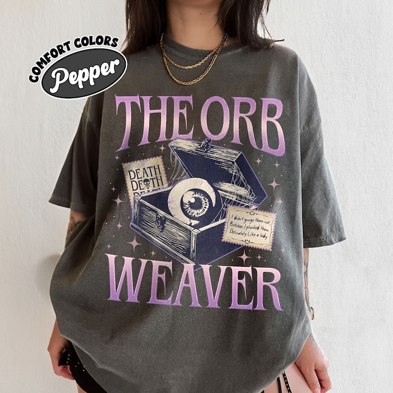 May include: A dark gray t-shirt with a graphic print of an eye inside a wooden box with the text "THE ORB WEAVER" in purple. The box has a spiderweb design and a label that reads "DEATH DEATH".