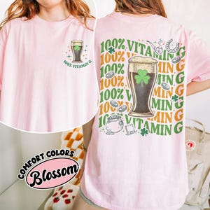 May include: A pale pink t-shirt featuring a beer glass with a shamrock and the text "100% Vitamin G". The back of the shirt has a larger graphic with the same theme, including a pot of gold and a horseshoe.