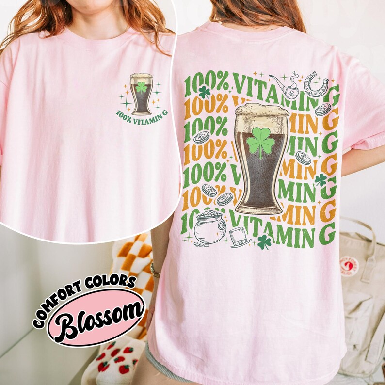 May include: A pale pink t-shirt featuring a beer glass with a shamrock and the text "100% Vitamin G". The back of the shirt has a larger graphic with the same theme, including a pot of gold and a horseshoe.