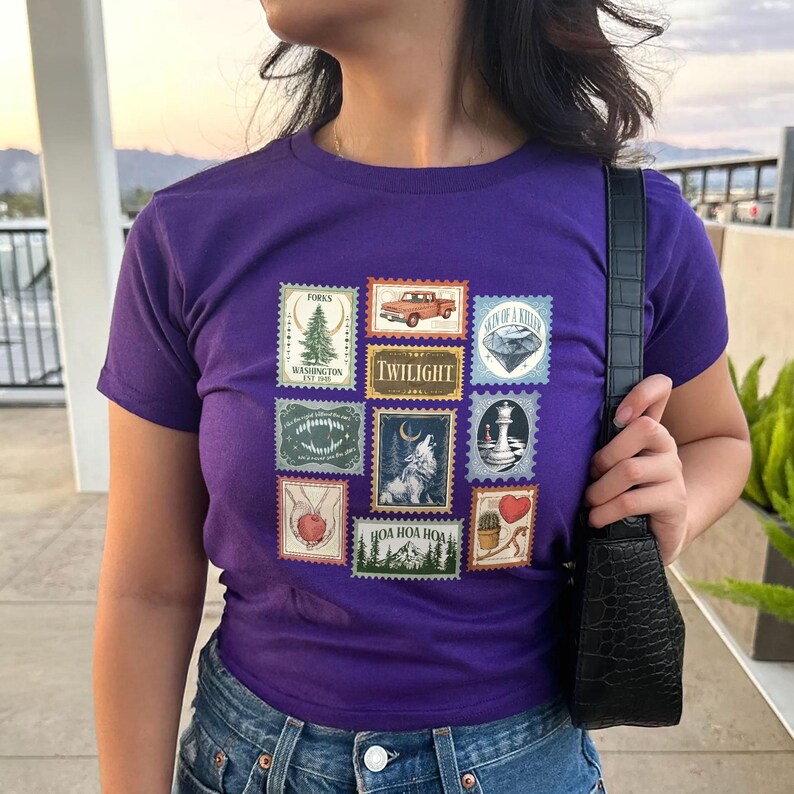 May include: A purple t-shirt featuring a collection of postage stamp-style graphics. The stamps depict various images, including a tree, a truck, a diamond, a wolf, and the word "Twilight". The shirt has a crew neck and short sleeves.