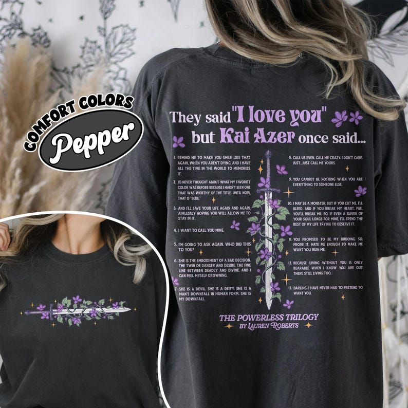 May include: Dark gray t-shirt with the text "They said 'I love you' but Kai Azer once said..." and a sword design with purple flowers. The shirt also features a list of quotes from the book "The Powerless Trilogy" by Lauren Roberts.