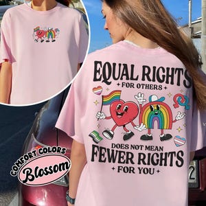 May include: Pink t-shirt with a graphic of two cartoon characters, one a heart and the other a rainbow, holding hands. The text "EQUAL RIGHTS + FOR OTHERS +" is above the characters and "DOES NOT MEAN FEWER RIGHTS + FOR YOU +" is below the characters.