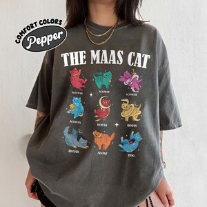Puede incluir: Una camiseta gris con el texto "THE MAAS CAT" impreso en la parte delantera. La camiseta presenta una cuadr&iacute;cula de nueve ilustraciones de gatos coloridos, cada uno con un dise&ntilde;o &uacute;nico y un nombre escrito debajo.