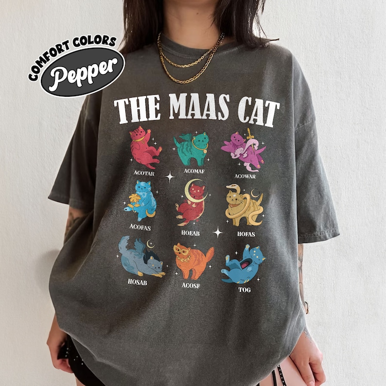 Puede incluir: Una camiseta gris con el texto "THE MAAS CAT" impreso en la parte delantera. La camiseta presenta una cuadr&iacute;cula de nueve ilustraciones de gatos coloridos, cada uno con un dise&ntilde;o &uacute;nico y un nombre escrito debajo.