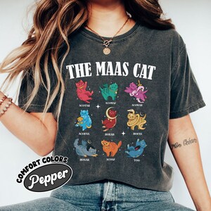 Puede incluir: Una camiseta gris con el texto "THE MAAS CAT" impreso en la parte delantera. La camiseta presenta una cuadr&iacute;cula de nueve gatos de dibujos animados coloridos con diferentes nombres impresos debajo de cada gato. Los nombres son: ACOTAR, ACOMAF, ACOWAR, ACOFAS, HOEAB, HOFAS, HOSAB, ACOSF y TOG.