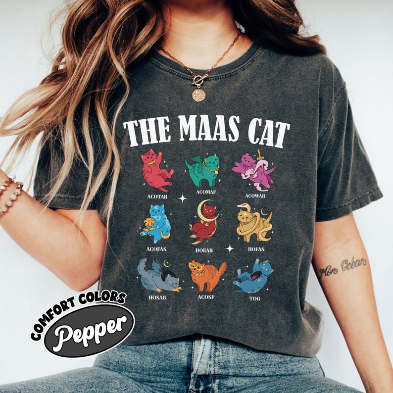 Puede incluir: Una camiseta gris con el texto "THE MAAS CAT" impreso en la parte delantera. La camiseta presenta una cuadr&iacute;cula de nueve gatos de dibujos animados coloridos con diferentes nombres impresos debajo de cada gato. Los nombres son: ACOTAR, ACOMAF, ACOWAR, ACOFAS, HOEAB, HOFAS, HOSAB, ACOSF y TOG.