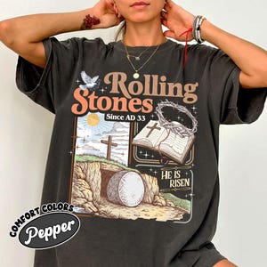 May include: Dark gray t-shirt with a graphic design featuring the text "Rolling Stones Since AD 33". The design includes a dove, a cross, an open book, and the phrase "He Is Risen". The shirt has a vintage, distressed look.