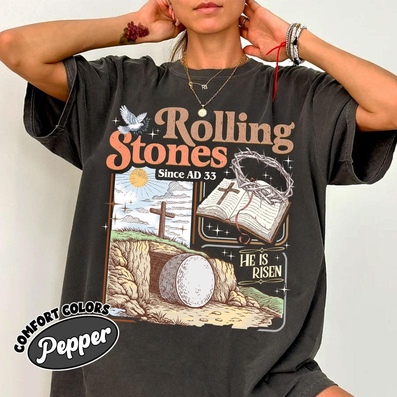 May include: Dark gray t-shirt with a graphic design featuring the text "Rolling Stones Since AD 33". The design includes a dove, a cross, an open book, and the phrase "He Is Risen". The shirt has a vintage, distressed look.
