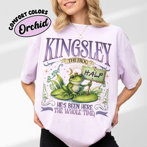 Puede incluir: Camiseta Orchid Comfort Colors con un dise&ntilde;o caprichoso. El dise&ntilde;o incluye el texto "KINGSLEY THE FROG" sobre una ilustraci&oacute;n de una rana sosteniendo un letrero que dice "HALP". El texto "HE'S BEEN HERE THE WHOLE TIME" est&aacute; debajo.