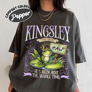 Puede incluir: Camiseta Comfort Colors gris oscuro con un gr&aacute;fico de una rana llamada Kingsley sosteniendo un letrero que dice "HALP". La camiseta tambi&eacute;n presenta el texto "He's Been Here The Whole Time!" y tiene un corte relajado.