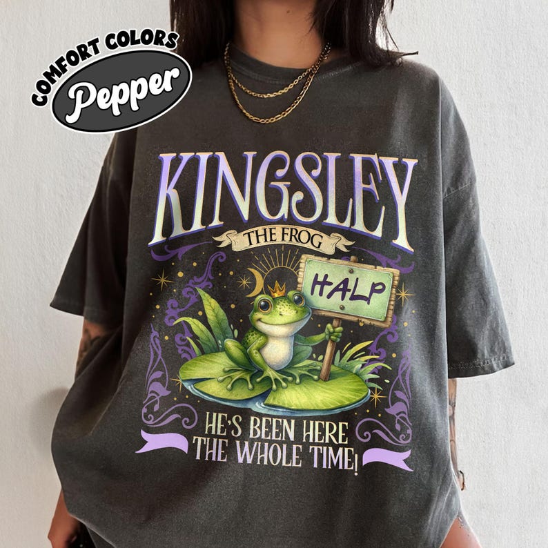 Puede incluir: Camiseta Comfort Colors gris oscuro con un gr&aacute;fico de una rana llamada Kingsley sosteniendo un letrero que dice "HALP". La camiseta tambi&eacute;n presenta el texto "He's Been Here The Whole Time!" y tiene un corte relajado.