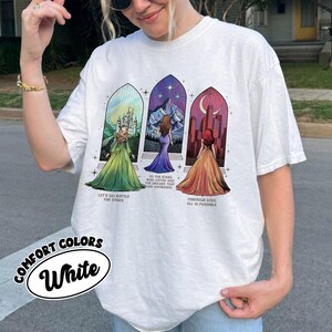 May include: White t-shirt with a colorful graphic print of three women in dresses standing in front of stained glass windows. The text "Let's go battle the stars", "To the stars who listen and the dreams that are answered", and "Through love all is possible" is printed below the women.