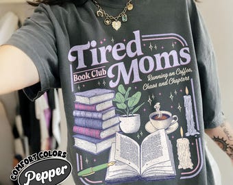 Può includere: T-shirt grigio scuro con la scritta "Tired Moms Book Club" in un design viola e bianco. La grafica include illustrazioni di libri, una tazza di caffè, candele e una pianta. La maglietta ha anche il testo "Running on Coffee, Chaos and Chapters."